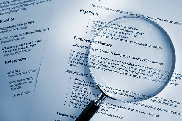 A blue tinted image of a magnifying glass lying on top of several sheets of a printout of resume documents. All information on the resume is fake.